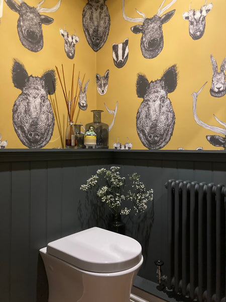 A wall adorned with a mustard yellow wallpaper featuring a pattern of hand-drawn British animals such as deer and a wild boar, with a vase of dried flowers in the foreground.